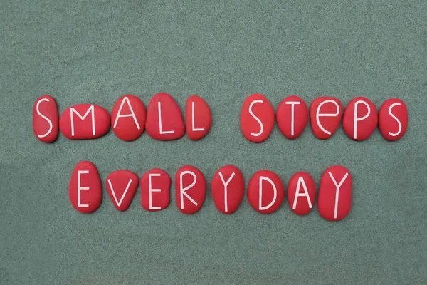 Her gün küçük adımlar, yeşil kum üzerinde kırmızı renkli taş harflerle oluşturulan motivasyon cümlesi.