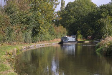 Güneşli bir günde kanala bakan gri bir dar kayık, sahil yoluna demirlemiş ve durgun suya yansıyor.