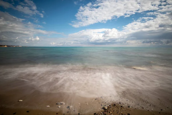Zeegolven spatten op de zandkust met kiezelsteentjes en blauwe bewolkte lucht. — Stockfoto