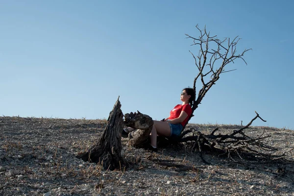 若い女性は 砂漠の丘の乾燥した木のログに座っている 乾いた土地に水はない ニコシアキプロス — ストック写真