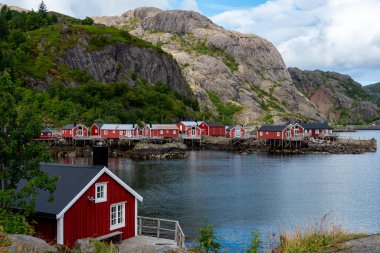 Sakin kayalık kıyı şeridi boyunca ayaklıklar üzerinde canlı kırmızı kulübeler yığını duruyor. Nusfjord balıkçı köyü Lofoten Adaları Norveç