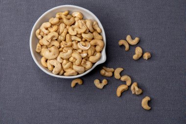 Kavrulmuş kaju fıstığıyla dolu beyaz seramik bir kase. Gri bir yüzeye yerleştirilmiş. Doğal ve iştah açıcı bir görünüş için etrafa saçılmış birkaç fındık..