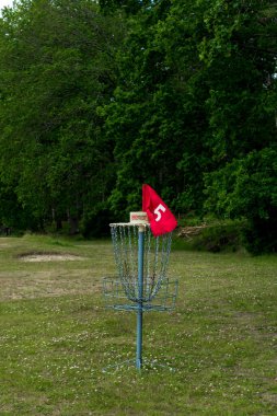 Parktaki yemyeşil ağaçlarla çevrili çimenli bir arazide duran zincirli ve kırmızı işaretli metal bir disk golf sepeti..