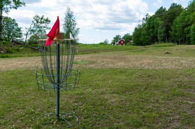 Parktaki yemyeşil ağaçlarla çevrili çimenli bir arazide duran zincirli ve kırmızı işaretli metal bir disk golf sepeti..