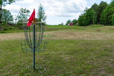 Parktaki yemyeşil ağaçlarla çevrili çimenli bir arazide duran zincirli ve kırmızı işaretli metal bir disk golf sepeti..