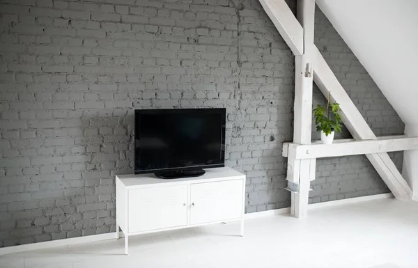 Modern flat lcd television set on white cabinet. Grey brick wall and wooden ceiling beams. Modern, bright attic apartment.