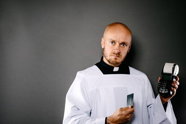 Priest in white surplice and black shirt with white collar holding credit card terminal. Church and money concept