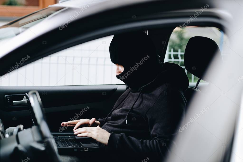 Masked thief hacker in a balaclava disarming car security systems and stealing a car