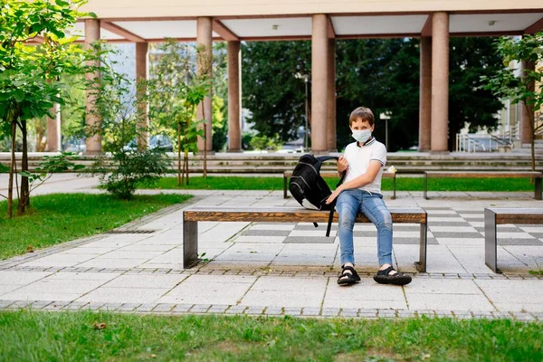 Um estudante mascarado com a mochila da escola no banco. Escola durante a pandemia do coronavírus covid-19 — Fotografia de Stock