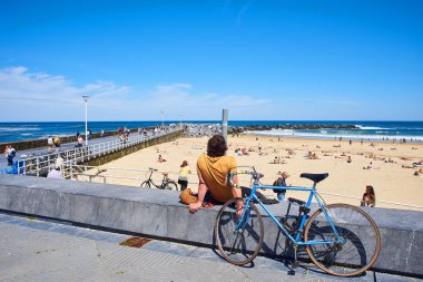 San Sebastian, Spain - May 11, 2018. A young men with their bike parked in front of  Zurriola beach and Monte Ulia in background at sunny day. San Sebastian, Guipuzcoa. Spain. clipart