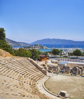 Bodrum, Turkey - July 6, 2018. Tourists visiting the Amphitheatre of Halicarnassus with Kumbahce bay and the Castle of Saint Peter in the background. Bodrum, Mugla Province, Turkey. clipart