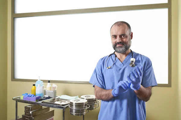 A Nurse putting on blue latex gloves in front of a nursing cart and a white window background.