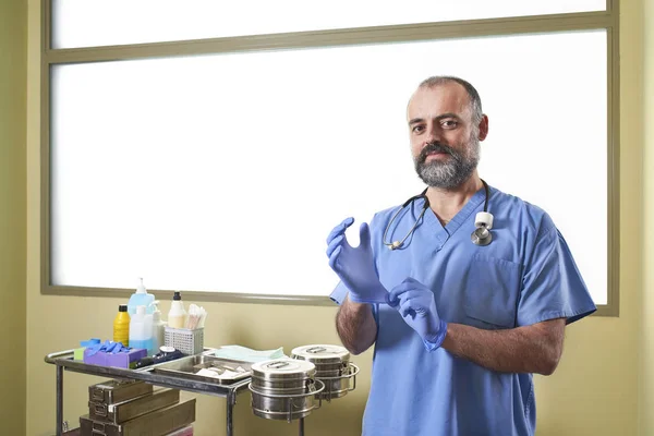 A Nurse putting on blue latex gloves in front of a nursing cart and a white window background.