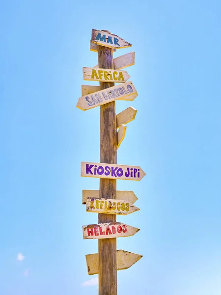 Tall post with colored wooden signboards pointing to various destinations and a beach services in Spanish.