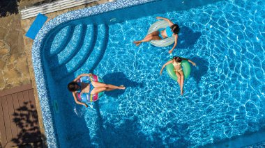 Yüzme havuzundaki aile yukarıdan insansız hava aracı görüntüsü, mutlu anne ve çocuklar şişme halka çöreklerle yüzüyor ve aile tatilinde suda eğleniyor, tatil beldesinde tropikal tatiller