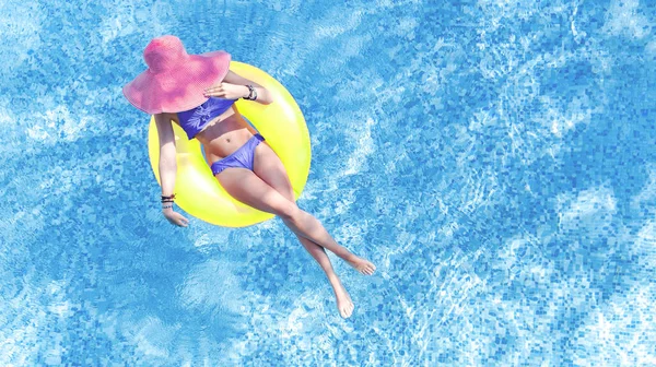 Beautiful young woman in hat in swimming pool aerial top view from above, young girl in bikini relaxes and swims on inflatable ring donut and has fun in water on family vacation, tropical holiday resort