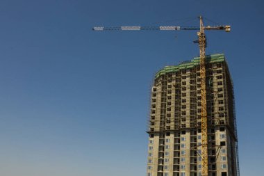 Büyük, yüksek beton bir ev, birçok kat, iskelenin etrafında. Büyük bir kule vincinin yanında. İnşaat endüstrisi, konut teslimatı, ortak inşaat. Mavi arkaplan, boşluğu kopyala.