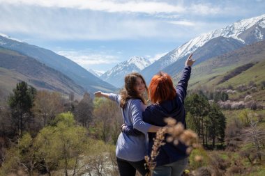 İki kız mutlu bir şekilde ellerini kaldırır. Güzel bir dağ geçidinin arka planında gezginler. Yüksek karlı dağlar ve önlerinde yeşil ağaçlar.