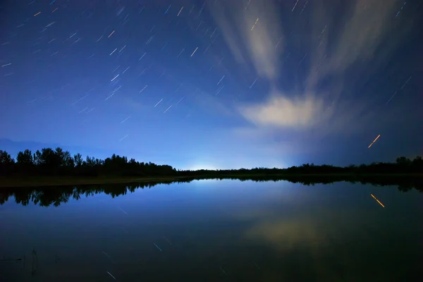 Starry sky and clouds. Night landscape, reflection of star paths in calm lake water.