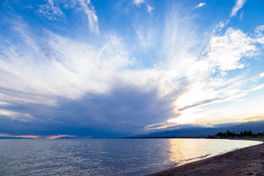Gölün kenarında güzel bir gün batımı. Parlak bulutlar suya yansıyor. Doğada harika bir yaz akşamı. Kırgızistan.