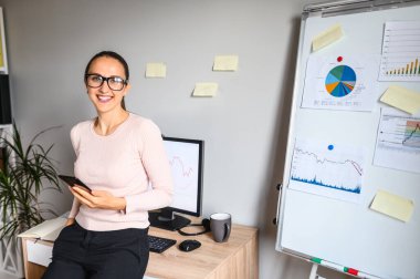 Gözlüklü, gülümseyen genç bir kadın elinde telefonla duruyor.