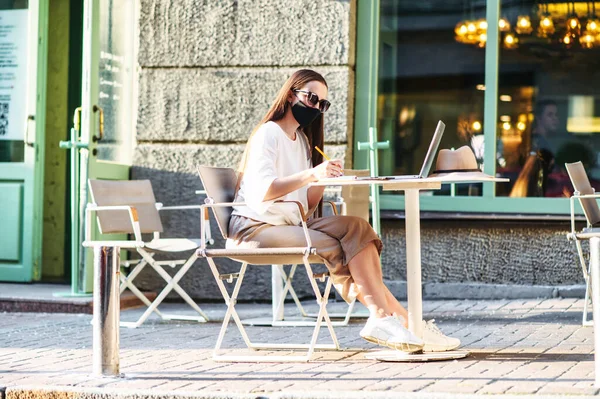 Жінка в медичній масці з ноутбуком в літньому кафе — стокове фото