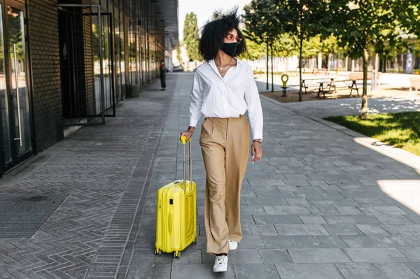 Dışarıda bavulu olan maskeli bir kadın.