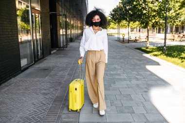 Dışarıda bavulu olan maskeli bir kadın.