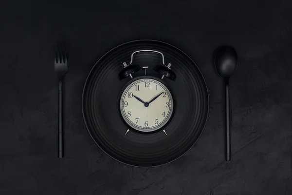 Black Alarm clock on black plate, spoon and fork on black background.