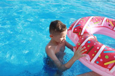 Güneşli bir günde havuzda, şişme yatakta pembe bir çörek şeklinde mutlu bir çocuk.