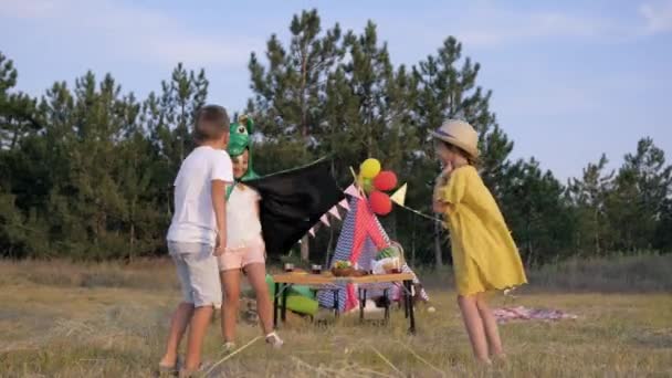 Aktív gyermekek, örömteli kislány a karneváli jelmezben fut a barátaival játszó energikus játék rét az erdőben során piknik a háttérben Wigwam — Stock videók