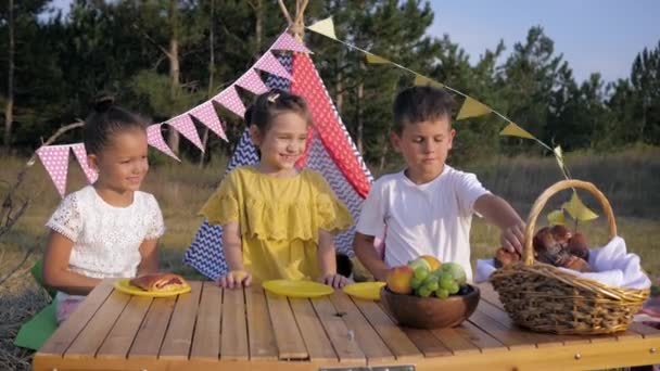 Dzieci związek, mały chłopiec dba o dziewcząt traktuje je do wypieków i jedzą traktuje na piknik w wiejskiej okolicy podczas słonecznych wakacji na tle Wigwam — Wideo stockowe