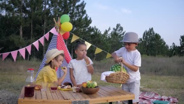 Śmieszne dzieci, mały chłopiec przynosi arbuz do kobiet przyjaciół podczas pikniku na imprezie leśnej na tle Wigwam — Wideo stockowe