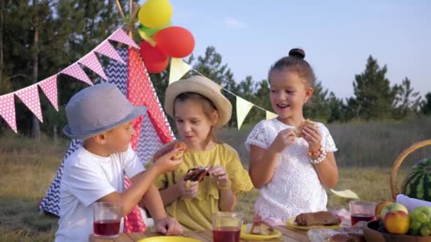 Kleine jongen feeds op gebak naar mooie vriendin tijdens het picknicken op het bos gazon terwijl ontspannen op het platteland achtergrond van Wigwam — Stockvideo