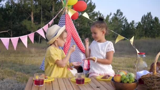 Μικρές φιλενάδες παίζουν παιχνίδι με ψαλίδι χαρτί ροκ και χυμό ποτού και κατά τη διάρκεια σνακ για πικ-νικ στις καλοκαιρινές διακοπές με φόντο την καλυβοσκηνή — Αρχείο Βίντεο