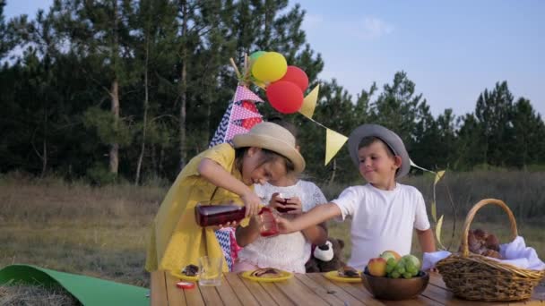 Pequeños en el picnic, chica encantadora vierte jugo de fruta en vasos a los amigos durante el almuerzo al aire libre en el bosque contra el telón de fondo de wigwam — Vídeos de Stock