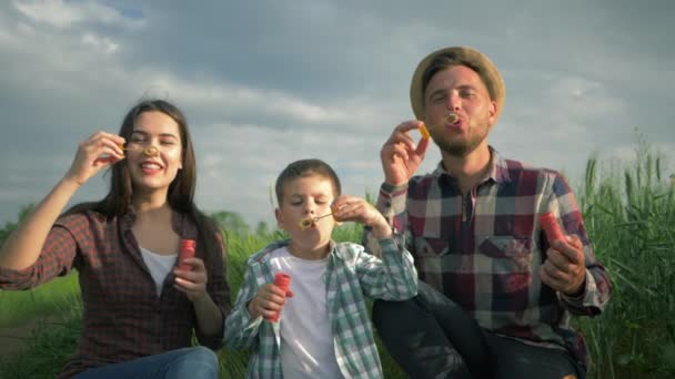 Bela jovem família soprada grandes bolhas em câmera lenta, pais com filho em camisas xadrez se divertir ao ar livre sentado no campo verde no fundo do céu — Vídeo de Stock