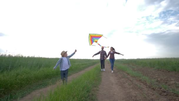Ребенок с воздушным змеем в руках бежит рядом с молодыми родителями в сельской местности в замедленном движении на фоне солнечного неба в выходные дни на открытом воздухе — стоковое видео