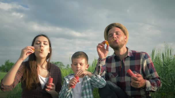 Familie pustet Seifenblasen und lacht in Zeitlupe, kleiner Junge mit Mama und Papa in karierten Hemden amüsieren sich am Wochenende auf der Wiese — Stockvideo