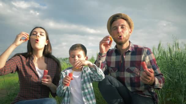 Portret młodej rodziny bawią się bańki mydlane na zewnątrz, dziecko z mamą i tatą w kratkę koszulki mają czas zabawy w polu na tle nieba — Wideo stockowe