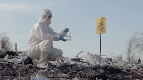 Wissenschaftlerin in Uniform und Schutzhandschuhen mit Reagenzgläsern, die Abfallproben von der großen Mülldeponie in der Nähe eines Hinweisschildes nehmen — Stockvideo