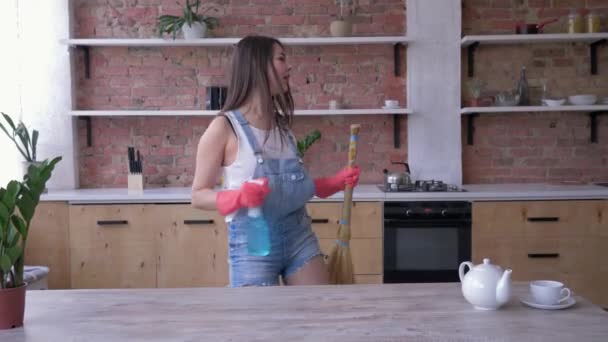 Nettoyage de cuisine heureux, portrait de jeune femme au foyer en salopette de jean et gants dansent heureux avec balai et spray nettoyant dans ses mains — Video