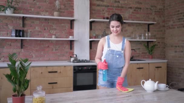 Ama de casa en overoles de mezclilla y guantes con trapo y spray de limpieza en las manos posando para la cámara en la cocina — Vídeos de Stock