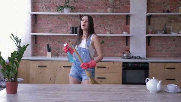 Spring cleaning, smiling girl in denim overalls and gloves happily has fun in kitchen with a broom and cleanser spray in her hands — стоковое видео