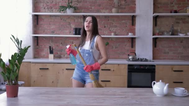 Lente reiniging, glimlachend meisje in denim overalls en handschoenen gelukkig heeft plezier in de keuken met een bezem en Cleanser spray in haar handen — Stockvideo