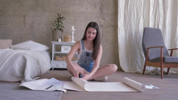 Reparação e quarto interior, menina sorridente em macacão denim medindo papel de parede com fita métrica no chão em plano — Vídeo de Stock