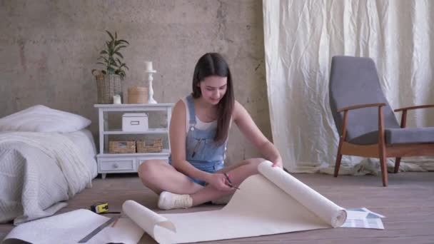 Reparatie appartement, jonge vrouw in denim overalls bezuinigingen nieuwe behang roll met schaar dan bekijken met glimlach op een cut stuk zittend op de vloer indoor — Stockvideo