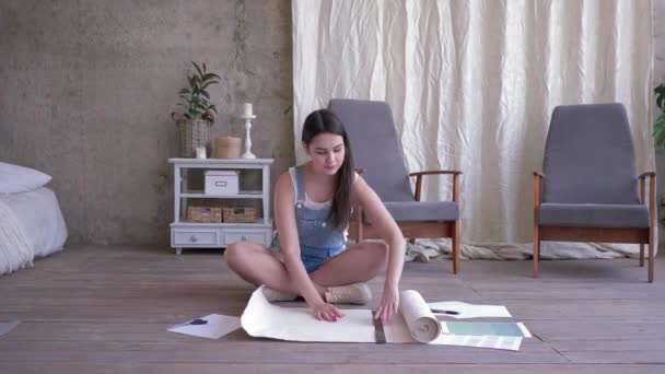 Chica joven desenrollando pedazo de papel pintado blanco hace marcas de lápiz y luego corta pieza con tijeras sentadas en el suelo en una habitación nueva — Vídeos de Stock