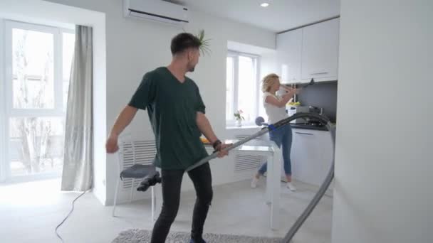 Беззаботная пара валяет дурака на кухне, мужчина танцует с пылесосом с девушкой, которая поет с ковш, как микрофон — стоковое видео