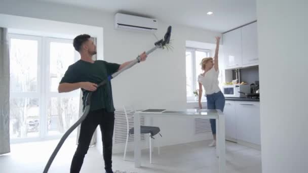 Веселая пара валяет дурака на кухне, парень танцует с пылесосом с девушкой, которая поет ложкой, как микрофон — стоковое видео